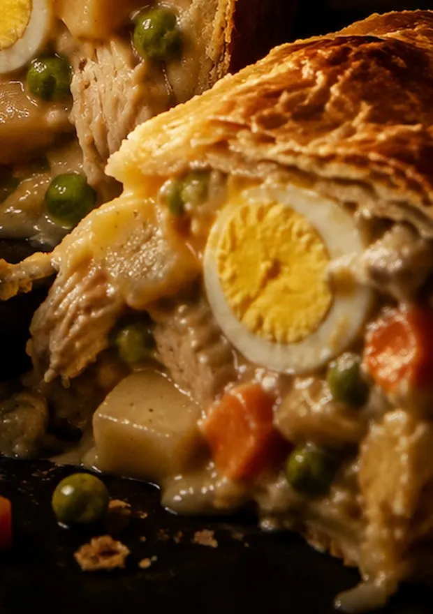 A slice of chicken pie with a flaky lid over creamy filling, half an egg, potato and peas