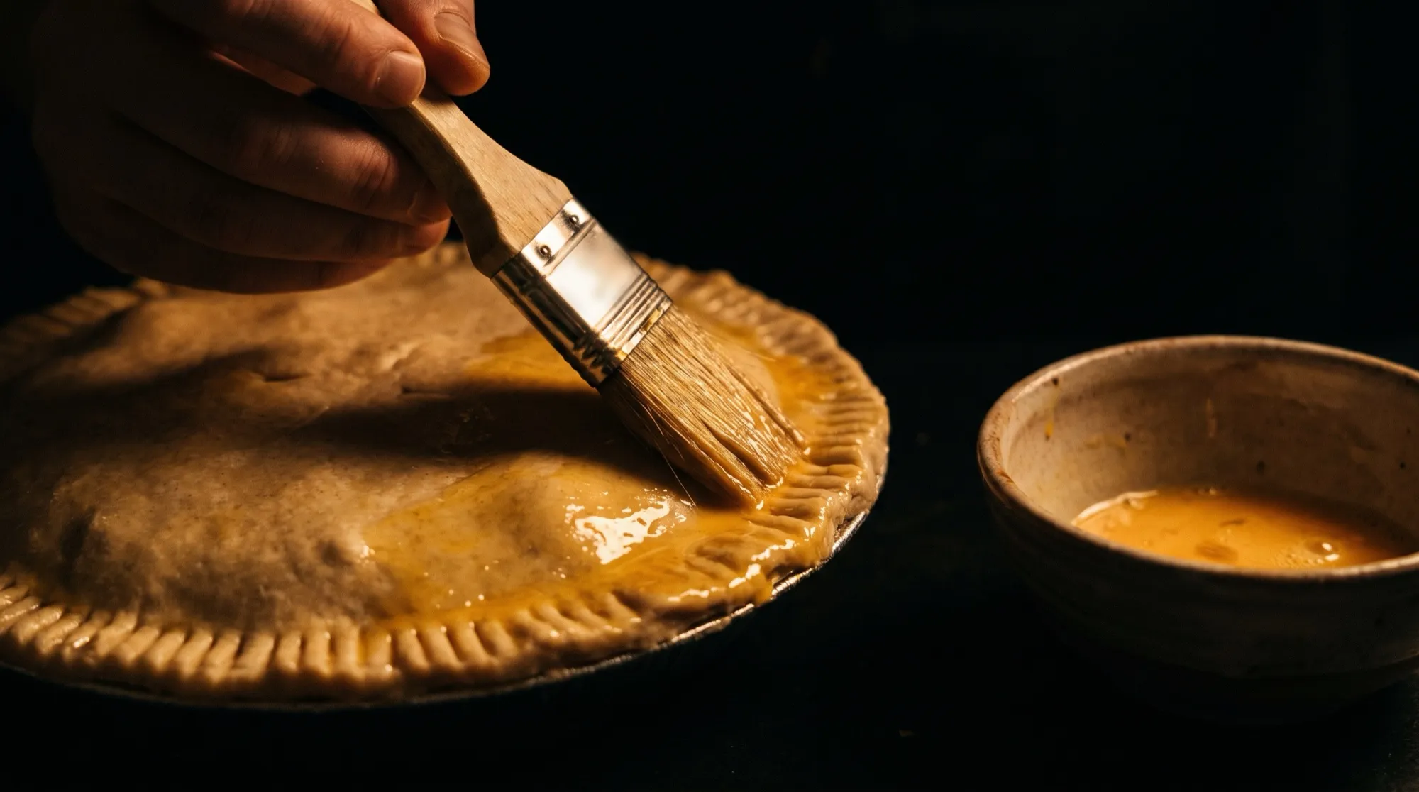 A pastry brush spreading glossy egg wash over the crimped lid of an unbaked chicken pie, a bowl of beaten egg beside it